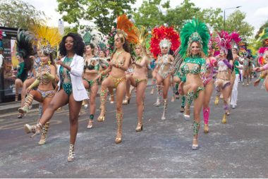 Carnival 2022 - Samba dancers in feathered costumes with knees up and in formation, the leader wears a white jacket. All the dancers are smiling.