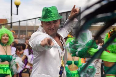 Carnival 2022 - Man in green hat pointing at camera, there are black feathers on the right of the view and bright green carnival outfits worn by dancers behind him